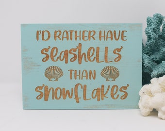 May include: A light blue wooden sign with the text "I'd rather have seashells than snowflakes" in brown lettering. Two seashells are also carved into the sign.