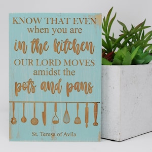 May include: A light blue wooden sign with a white distressed background. The sign has a quote from St. Teresa of Avila: "Know that even when you are in the kitchen our Lord moves amidst the pots and pans." The sign has a row of kitchen utensils hanging from the top.