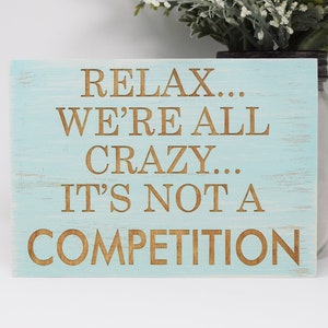 May include: A light blue wooden sign with the text "Relax... We're all crazy... It's not a competition" in brown lettering.