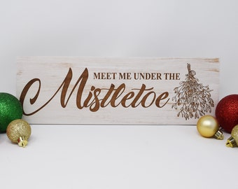 May include: White wooden sign with the text "Meet me under the mistletoe" in brown lettering. The sign is decorated with a brown illustration of mistletoe. The sign is surrounded by gold and green Christmas ornaments.