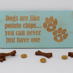 May include: A light blue wooden sign with the text "Dogs are like potato chips... you can never just have one" in brown lettering. Two brown paw prints with hearts are on the right side of the sign.