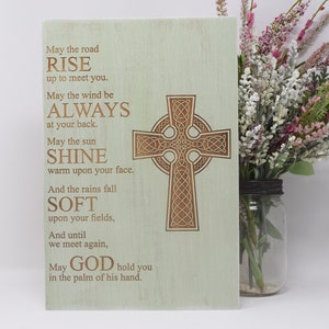 Puede incluir: Placa de madera verde claro con un diseño de cruz celta y una bendición irlandesa. El texto incluye frases como "Que el camino se alce para encontrarte" y "Que Dios te sostenga en la palma de su mano."