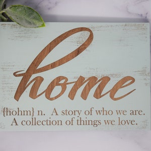 May include: A wooden sign with a distressed light blue background and brown lettering. The sign says "home" in a cursive font and includes the definition: "{hohm} n. A story of who we are. A collection of things we love."