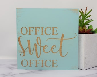May include: A square, light blue wooden sign with the words "OFFICE Sweet OFFICE" carved into the wood in a cursive font. The sign has a distressed look with a light brown color. A white pot with a succulent plant is to the right.