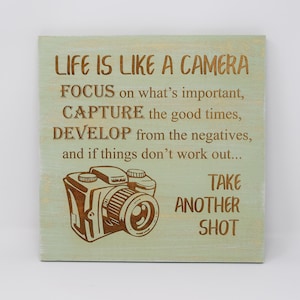 May include: A wooden sign with a green background and a brown camera illustration. The sign has the text "LIFE IS LIKE A CAMERA FOCUS on what's important, CAPTURE the good times, DEVELOP from the negatives, and if things don't work out... TAKE ANOTHER SHOT".