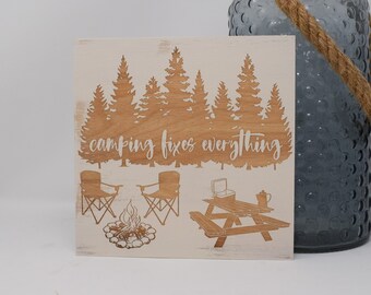 May include: White wooden sign with a woodgrain texture featuring a camping scene with trees, a campfire, two chairs, a picnic table, a cooler, and a coffee pot. The text "camping fixes everything" is written in white on the sign.