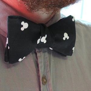 May include: A black bow tie with a white Mickey Mouse pattern is fastened around a person's neck. The bow tie is made of fabric and is paired with a gray button-down shirt. The background is a soft pink.