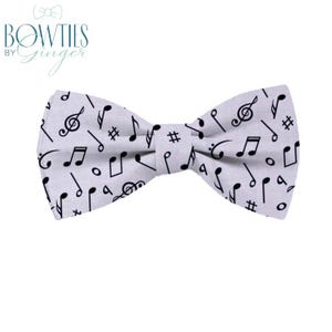 May include: A light gray bow tie featuring a pattern of black musical notes, including treble clefs, eighth notes, and sharps. The bow tie is a classic accessory, perfect for formal events or adding a touch of musical flair to an outfit. The text "Bowties by Ginger" is in the upper left corner.