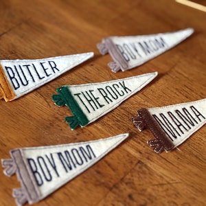May include: A collection of felt pennants with various text, including "BUTLER", "THE ROCK", "BOY MOM", and "MAMA". The pennants are white with dark blue lettering and colorful trim in shades of brown, green, and orange. They are arranged on a wooden surface.
