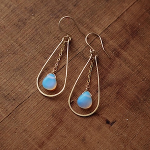 Puede incluir: Un par de pendientes dorados en forma de lágrima. Cada pendiente presenta una delicada cadena dorada que suspende una piedra preciosa azul claro en forma de pera. Los pendientes se exhiben sobre una superficie de madera.