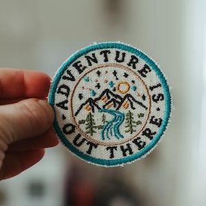 May include: Round embroidered patch with the text "Adventure is out there." The patch features a mountain range, trees, a river, and a sun design. The border is teal and the text is black.