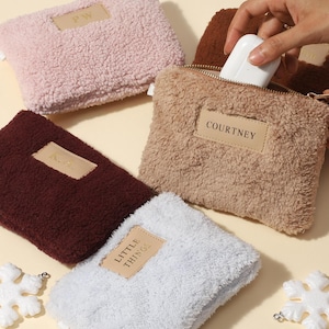 Könnte beinhalten: Eine Auswahl an flauschigen, strukturierten Pouches in verschiedenen Farben wie Rosa, Braun und Weiß. Jeder Pouch hat ein rechteckiges Lederetikett mit personalisiertem Text. Ein Pouch wird geöffnet, um ein weißes Objekt zu zeigen. Dekorative Schneeflocken-Ornamente sind verstreut.