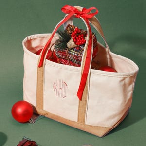 May include: A canvas tote bag with a monogram, tan trim, and red ribbon. The bag is filled with Christmas ornaments and a wrapped gift. A red, green, and gold ornament are scattered around the bag on a green background.