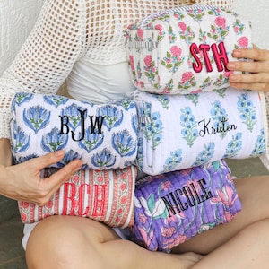 May include: A collection of personalized cosmetic bags in various floral patterns and colors. The bags feature embroidered initials or names in contrasting colors. The bags are in shades of pink, blue, red, and purple.
