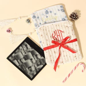 Puede incluir: Dos fundas de tela acolchada, una con estampado floral y el nombre "Emily", la otra con monograma y lazo rojo. Un lector digital, un bastón de caramelo, una piña y un adorno de copo de nieve completan el arreglo navideño.