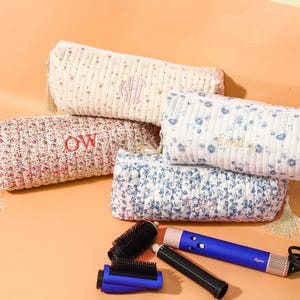 May include: Five floral print cosmetic bags in various colors, with embroidered initials. A blue and pink hair styling tool and accessories are also visible. The bags have tassel zipper pulls.
