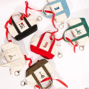 May include: A collection of small canvas tote bags in various colors, each with a unique design. The bags feature red, black, blue, and green accents, with embroidered details like letters, symbols, and the phrase "I ❤️ NY." Each bag has a red ribbon and a keychain.