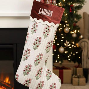 May include: A Christmas stocking with a claret cuff and the name "LAUREN" embroidered in white. The body of the stocking is white with a floral pattern in pink and green. The stocking hangs from a white mantelpiece.