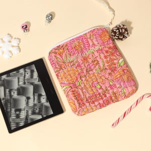 Puede incluir: Una bolsa con cremallera con estampado floral rosa, un lector de libros electr&oacute;nicos y adornos navide&ntilde;os est&aacute;n dispuestos sobre una superficie de color crema. La bolsa presenta un colorido estampado floral. Un bast&oacute;n de caramelo, una pi&ntilde;a y adornos se suman al tema festivo.