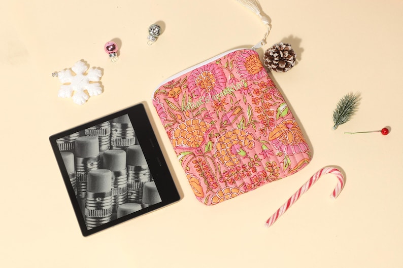 Puede incluir: Una bolsa con cremallera con estampado floral rosa, un lector de libros electr&oacute;nicos y adornos navide&ntilde;os est&aacute;n dispuestos sobre una superficie de color crema. La bolsa presenta un colorido estampado floral. Un bast&oacute;n de caramelo, una pi&ntilde;a y adornos se suman al tema festivo.