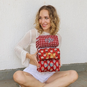May include: Three red cosmetic bags with embroidered initials. The top bag has a red and white checkered pattern with the initials "OFS". The middle bag has the initials "TM" and the bottom bag has the initials "AS".