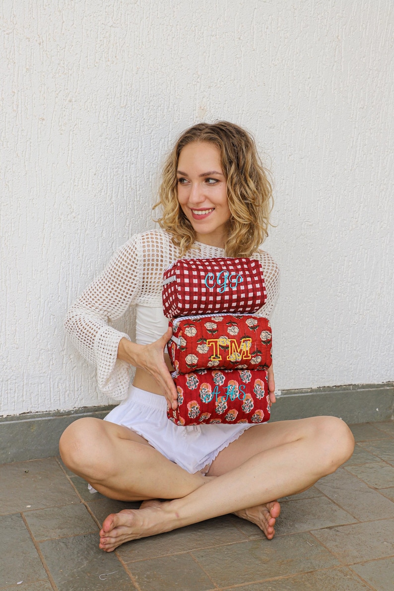 May include: Three red cosmetic bags with embroidered initials. The top bag has a red and white checkered pattern with the initials "OFS". The middle bag has the initials "TM" and the bottom bag has the initials "AS".