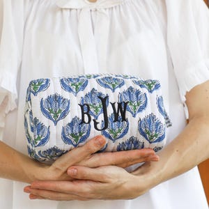 May include: A quilted cosmetic bag with a white background and a repeating blue floral pattern. The bag has a rectangular shape and features the embroidered initials "BJW" in black. The bag is held in front of a white blouse.