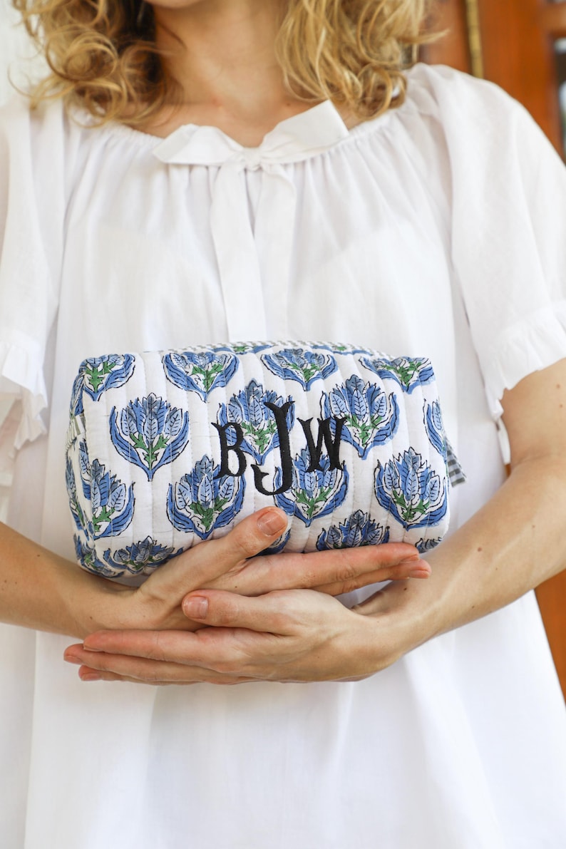 May include: A quilted cosmetic bag with a white background and a repeating blue floral pattern. The bag has a rectangular shape and features the embroidered initials "BJW" in black. The bag is held in front of a white blouse.