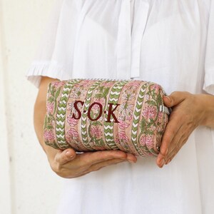 May include: A rectangular cosmetic bag with a floral pattern in pink and green, held by a person wearing a white top. The bag features the initials "S.O.K." in a dark red color.