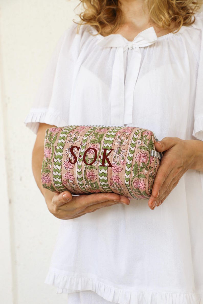 May include: A rectangular cosmetic bag with a floral pattern in pink and green, held by a person wearing a white top. The bag features the initials "S.O.K." in a dark red color.