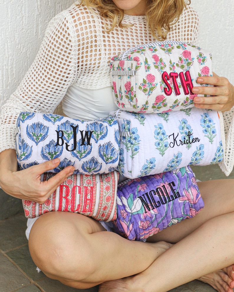 May include: A collection of personalized cosmetic bags in various floral patterns and colors. The bags feature embroidered initials or names in contrasting colors. The bags are in shades of pink, blue, red, and purple.