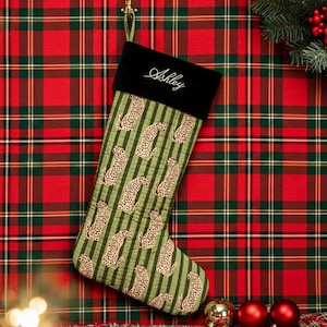 May include: A Christmas stocking with a black cuff embroidered with the name "Ashley" in white script. The body of the stocking is green with vertical stripes and leopard print. The background is a red and green plaid.