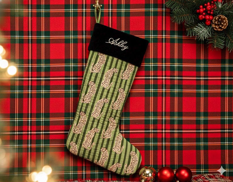 May include: A Christmas stocking with a black cuff embroidered with the name "Ashley" in white script. The body of the stocking is green with vertical stripes and leopard print. The background is a red and green plaid.