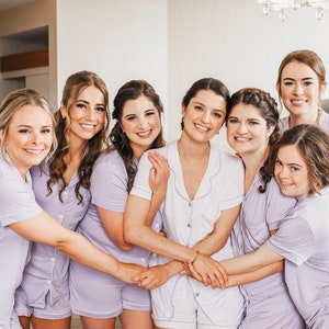 May include: A group of eight women wearing matching lavender pyjamas. They are smiling and posing for a photo. The women are all wearing short-sleeved pyjamas with a button-up front. The pyjamas have a white trim around the collar, cuffs, and pockets.
