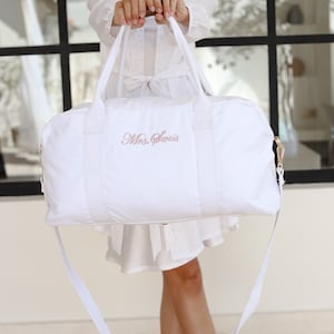 K&ouml;nnte beinhalten: Wei&szlig;e Reisetasche aus Canvas mit langem Riemen und einer rosa gestickten Aufschrift "Mrs. Sweiss".