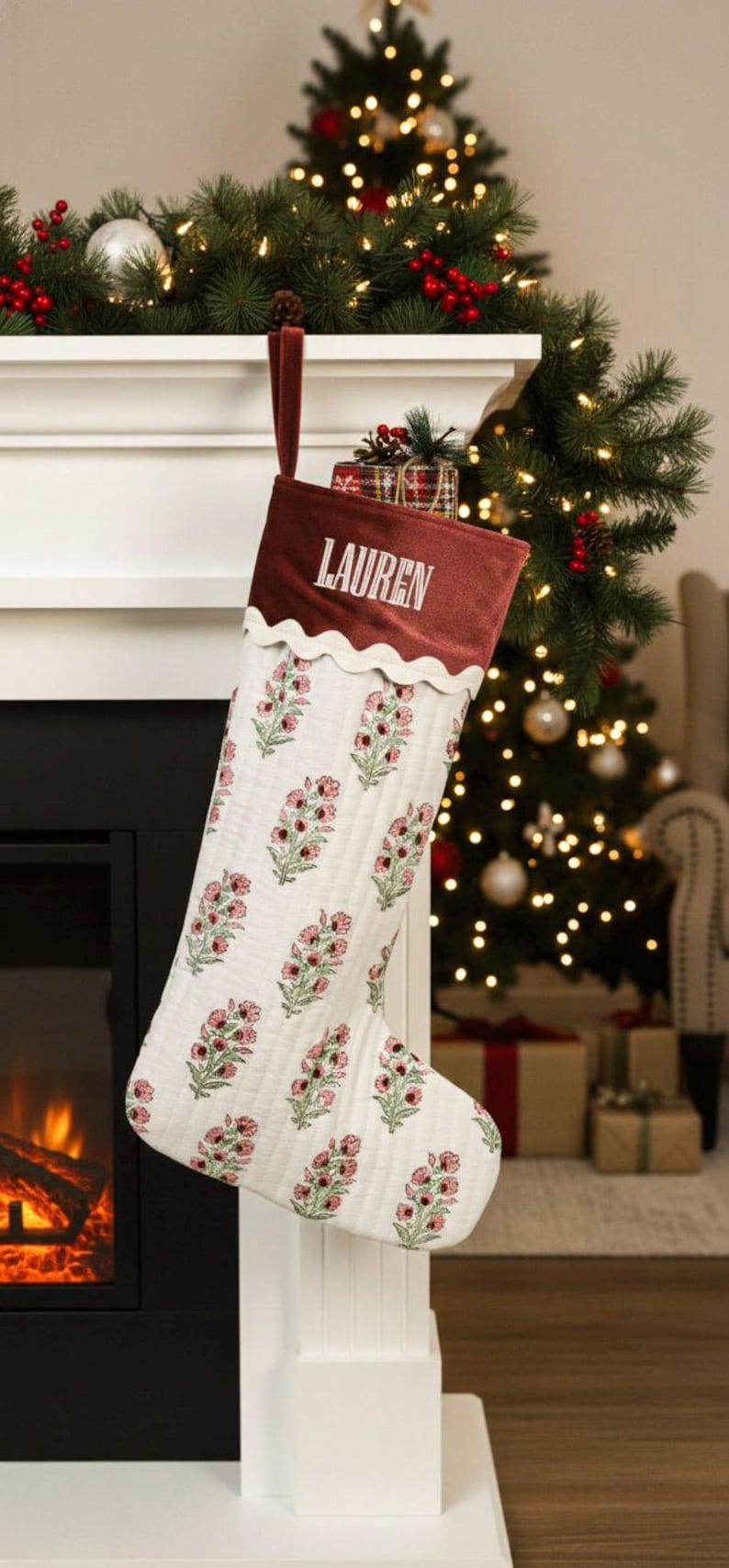 May include: A Christmas stocking with a burgundy cuff and the name "LAUREN" embroidered in white. The body of the stocking is white with a floral pattern in pink and green. The stocking hangs from a white mantel.