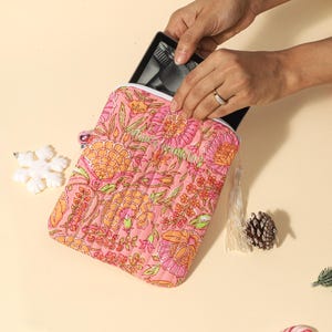 Puede incluir: Una funda acolchada para tableta de color rosa con estampado floral y detalles en naranja y verde. Se ve una tableta negra dentro. Una borla blanca y una pi&ntilde;a est&aacute;n al lado.