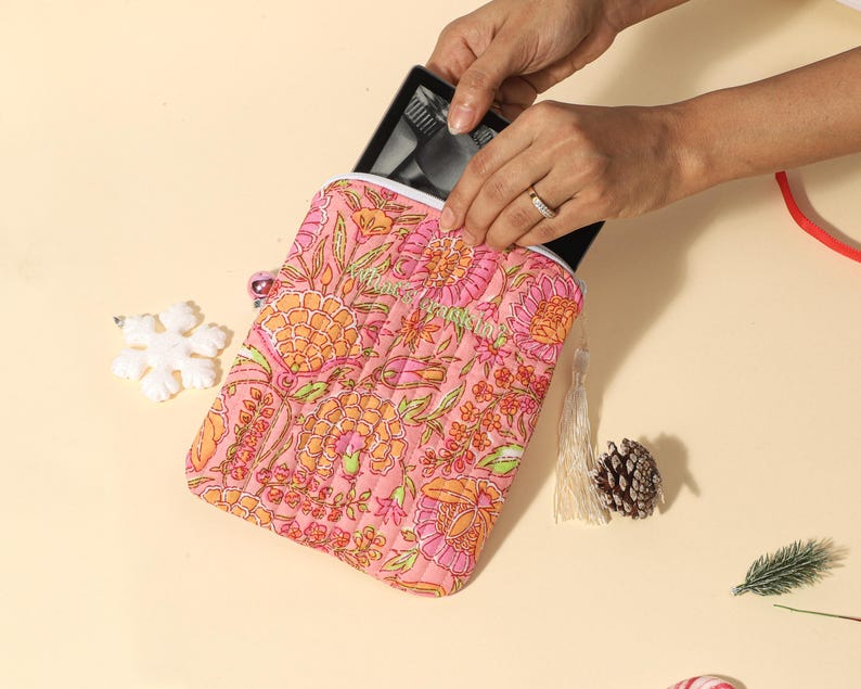 Puede incluir: Una funda acolchada para tableta de color rosa con estampado floral y detalles en naranja y verde. Se ve una tableta negra dentro. Una borla blanca y una pi&ntilde;a est&aacute;n al lado.