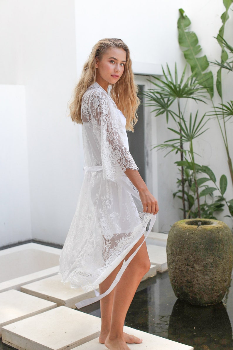 May include: A white lace robe with long sleeves and a tie belt. The robe is sheer and has a delicate floral pattern. The model is standing on a white tiled floor.