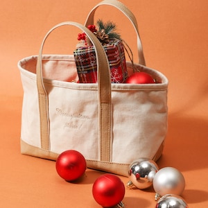 May include: A cream canvas tote bag with tan trim and handles, embroidered with "Cosmopolitan, please!". It contains a wrapped gift and red baubles. Silver and red baubles are scattered around the bag on an orange background.