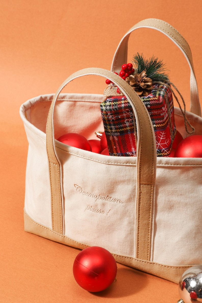 May include: A cream canvas tote bag with brown straps and base, containing red baubles and a wrapped gift. The gift is in a tartan pattern with a pine cone and berries. The bag has the text "Cosmopolitan please!"