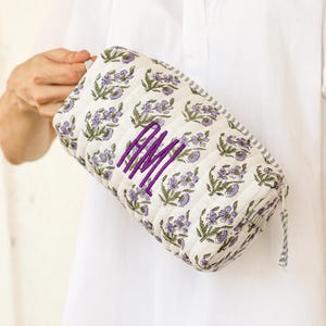 May include: A quilted cosmetic bag with a floral pattern in purple and green on a white background. The bag features a purple monogram and a striped trim. The bag is rectangular and has a zipper closure.