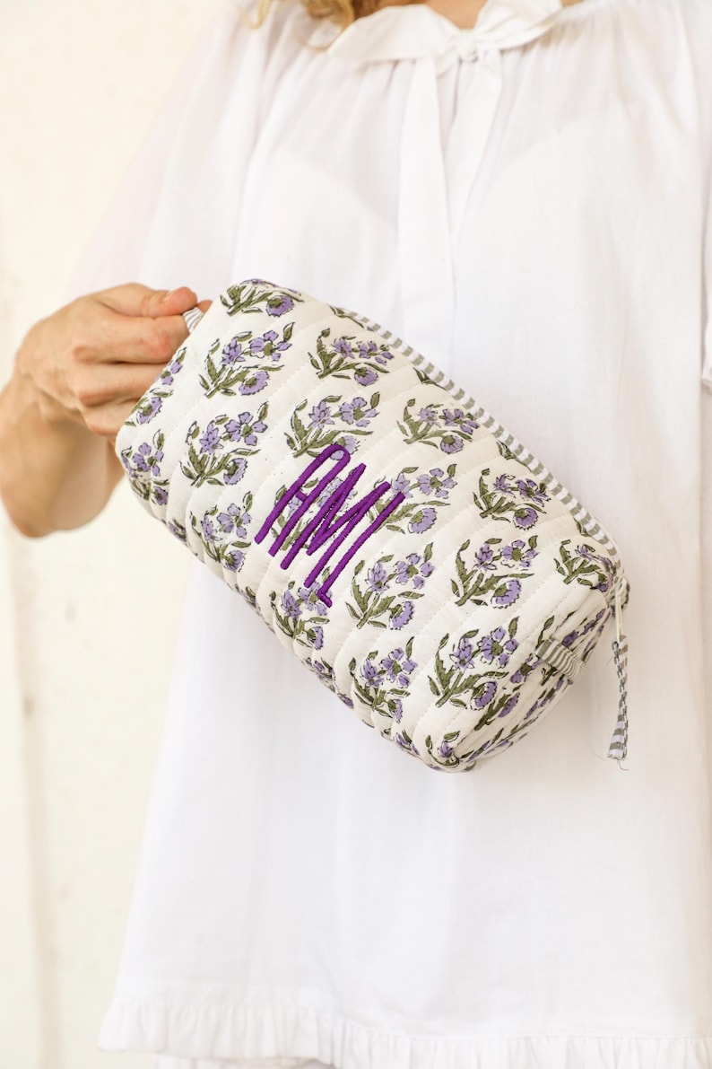 May include: A quilted cosmetic bag with a floral pattern in purple and green on a white background. The bag features a purple monogram and a striped trim. The bag is rectangular and has a zipper closure.