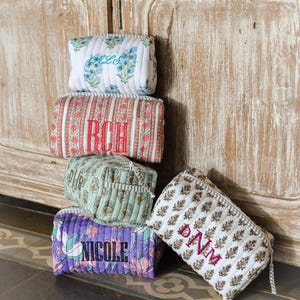 May include: A stack of quilted cosmetic bags in various floral and patterned designs. Each bag features embroidered initials or a name in contrasting colors. The bags are in shades of white, red, purple, green, and brown.