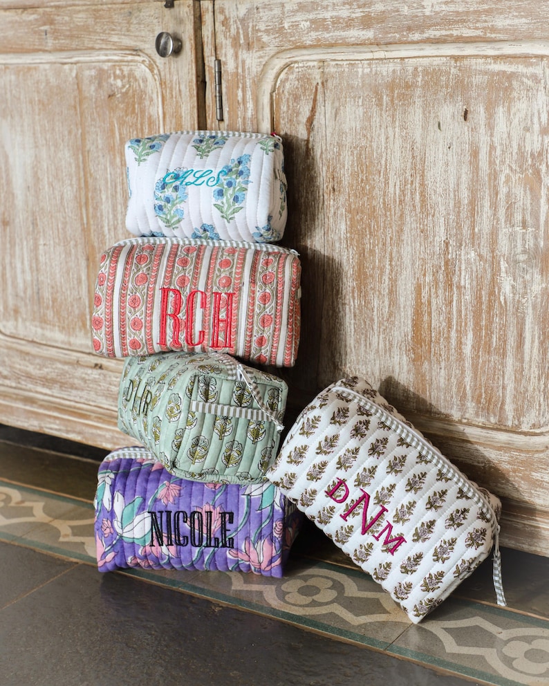 May include: A stack of quilted cosmetic bags in various floral and patterned designs. Each bag features embroidered initials or a name in contrasting colors. The bags are in shades of white, red, purple, green, and brown.