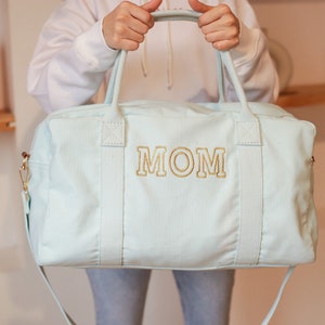 May include: A light blue canvas duffel bag with a gold embroidered "MOM" on the front. The bag has two top handles and a long shoulder strap.