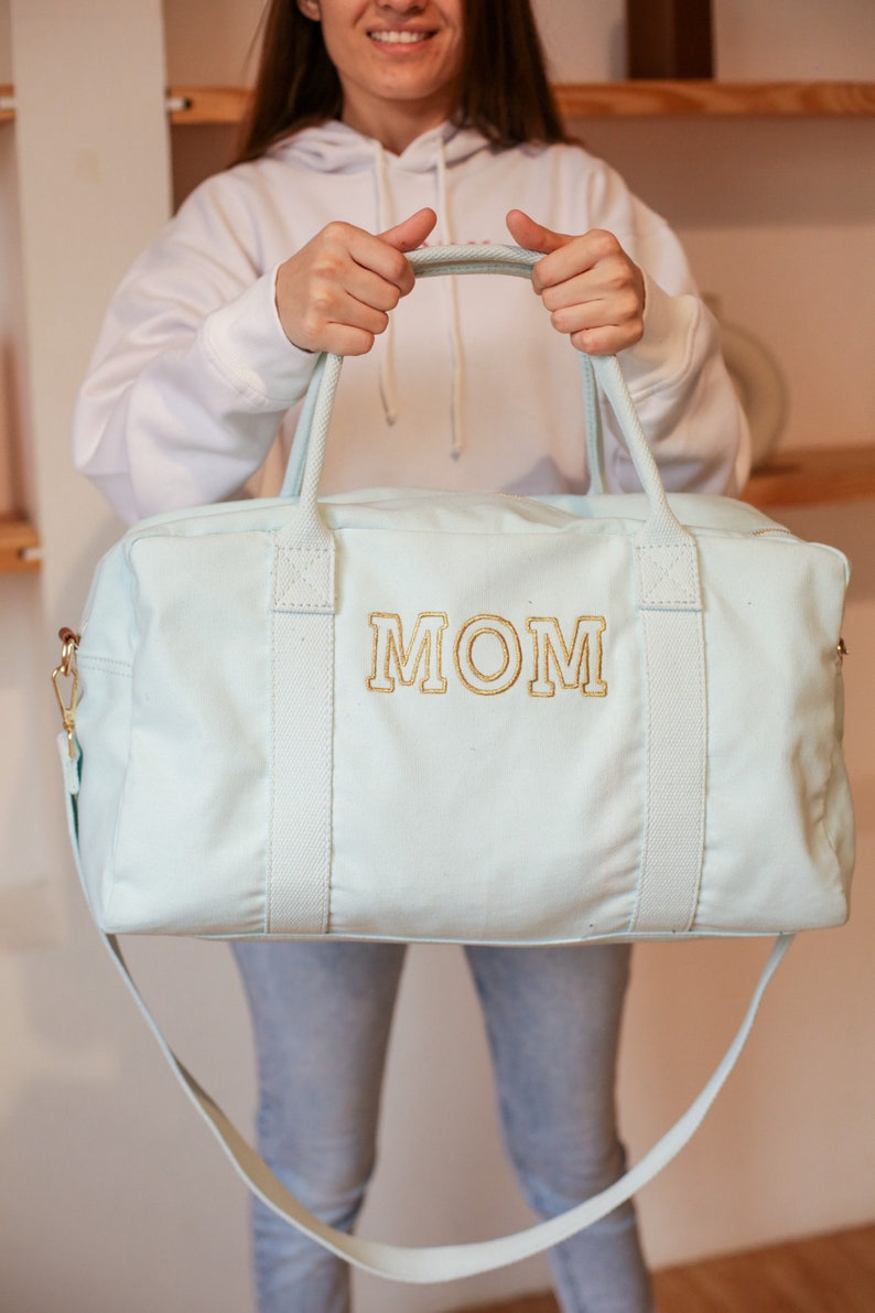 May include: A light blue canvas duffel bag with a gold embroidered "MOM" on the front. The bag has two top handles and a long shoulder strap.