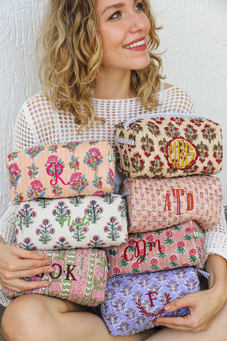 May include: A collection of colorful, floral-patterned cosmetic bags. Each bag features a unique embroidered initial or monogram in a contrasting color. The bags are in various shades of pink, peach, and lavender, with a quilted texture and a striped handle.
