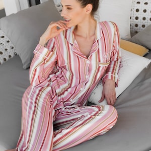 May include: A woman wearing pink and white striped pajamas sits on a white wicker couch. She is smiling and has her hand on her chin.