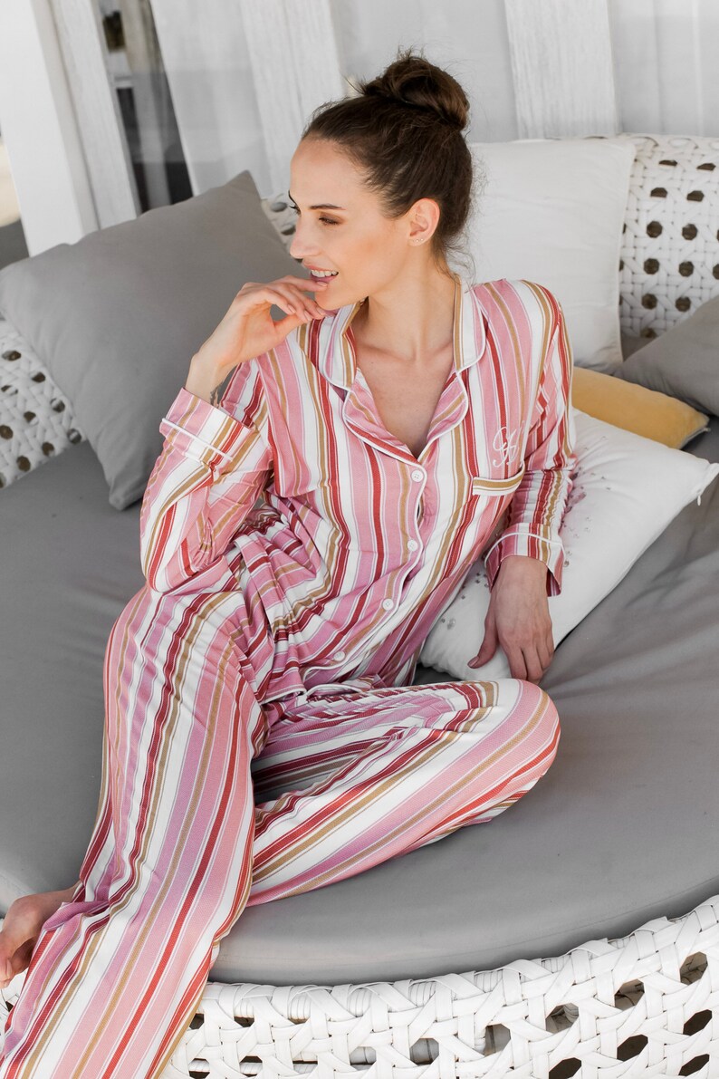 May include: A woman wearing pink and white striped pajamas sits on a white wicker couch. She is smiling and has her hand on her chin.